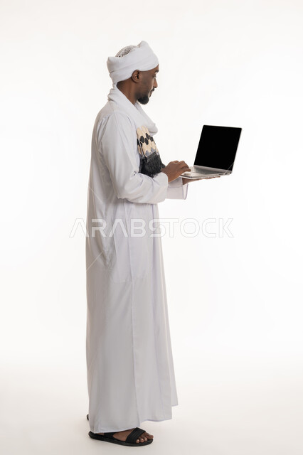 مفهوم إنجاز مهام العمل عن بعد, استخدام الأجهزة التقنية الحديثة والمتطورة, بورتريه من الجانب لرجل عربي سوداني يرتدي الجلابية والعمامة السودانية يقوم باستعمال جهاز الحاسوب المحمول, عرض شاشة سوداء فارغة عبر الحاسب, الجسم بالطول الكامل, خلفية بيضاء
