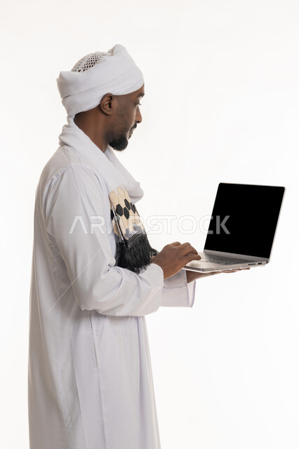 استخدام الأجهزة التقنية الحديثة والمتطورة, مفهوم إنجاز مهام العمل عن بعد, بورتريه من الجانب لرجل عربي سوداني يرتدي الجلابية والعمامة السودانية يقوم باستعمال جهاز الحاسوب المحمول, عرض شاشة سوداء فارغة عبر الحاسب, خلفية بيضاء