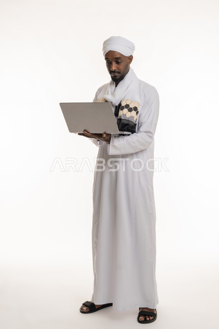 استخدام الأجهزة التقنية الحديثة والمتطورة, مفهوم إنجاز مهام العمل عن بعد, بورتريه لرجل عربي سوداني يرتدي الجلابية والعمامة السودانية يقوم باستعمال جهاز الحاسوب المحمول, الجسم بالطول الكامل, خلفية بيضاء