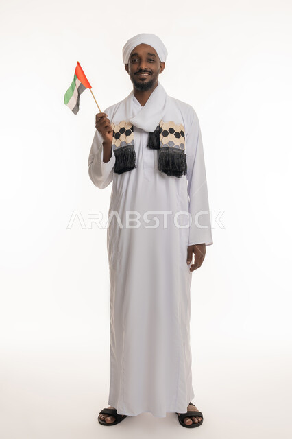 الاحتفال بالأعياد والمناسبات الوطنية, النظر إلى الكاميرا بإيماءات السعادة والسرور, اليوم الوطني الإماراتي 2 ديسمبر, استذكار يوم العلم 3 نوفمبر, بورتريه لرجل عربي سوداني يرتدي الجلابية والعمامة السودانية يحمل علم الإمارات, الجسم بالطول الكامل, خلفية بيضاء