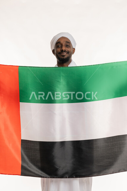Concept of love and belonging, celebrating national holidays and occasions, UAE National Day December 2, commemorating Flag Day November 3, portrait of a smiling Sudanese Arab man wearing a Sudanese galabiya and turban holding the flag of the United Arab Emirates, white background