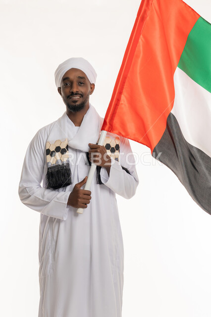 Looking at the camera with gestures of happiness and pleasure, celebrating national holidays and occasions, UAE National Day December 2, commemorating Flag Day November 3, portrait of a Sudanese Arab man wearing a Sudanese jalabiya and turban holding the flag of the United Arab Emirates, white background