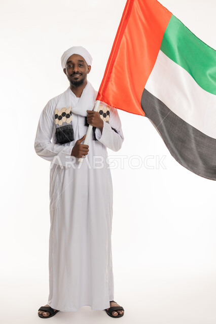 استذكار يوم العلم 3 نوفمبر, النظر إلى الكاميرا بإيماءات السعادة والسرور, الاحتفال بالأعياد والمناسبات الوطنية, اليوم الوطني الإماراتي 2 ديسمبر, بورتريه لرجل عربي سوداني يرتدي الجلابية والعمامة السودانية يحمل علم الإمارات, الجسم بالطول الكامل, خلفية بيضاء