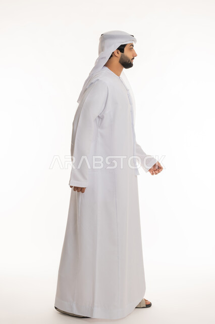 الذهاب إلى مكان ما, مفهوم الاهتمام بالأناقة والمظهر الخارجي, بورتريه من الجانب لرجل عربي خليجي إماراتي يرتدي الكندورة والغترة البيضاء يقوم بالمشي, الجسم بالطول الكامل, خلفية بيضاء