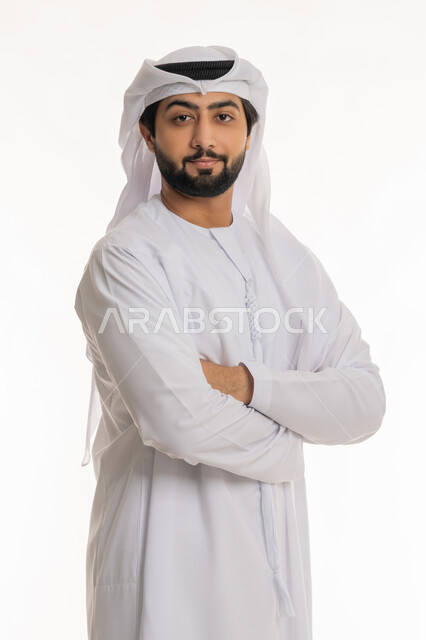 النظر إلى الكاميرا بإيماءات السعادة والسرور, مفهوم الأناقة والرجولة, الاهتمام بالمظهر الخارجي, بورتريه لرجل عربي خليجي إماراتي مبتسم يرتدي الكندورة والغترة البيضاء يقف بأيدي متقاطعة بتعابير الثقة بالنفس, خلفية بيضاء