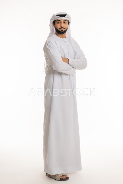 مفهوم الأناقة والرجولة, النظر إلى الكاميرا بإيماءات السعادة والسرور, الاهتمام بالمظهر الخارجي, بورتريه لرجل عربي خليجي إماراتي مبتسم يرتدي الكندورة والغترة البيضاء يقف بأيدي متقاطعة بتعابير الثقة بالنفس, الجسم بالطول الكامل, خلفية بيضاء
