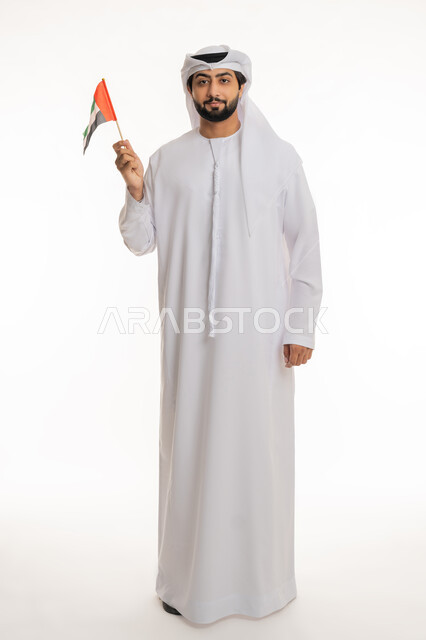 النظر إلى الكاميرا بإيماءات السعادة والسرور, مفهوم الحب والفخر بالوطن, ذكرى اليوم الوطني الإماراتي 2 ديسمبر, بورتريه لرجل عربي خليجي إماراتي يرتدي الكندورة والغترة البيضاء يحمل علم الإمارات بيده, الجسم بالطول الكامل، خلفية بيضاء