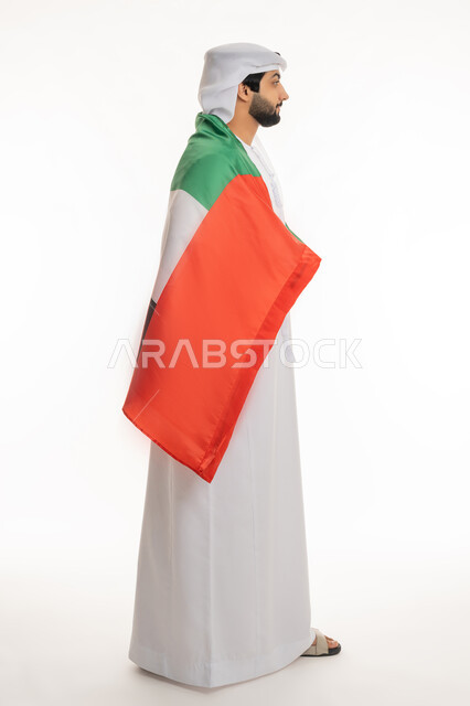 مفهوم الحب والفخر بالوطن, الاحتفال بالأعياد والمناسبات الوطنية, ذكرى اليوم الوطني الإماراتي 2 ديسمبر, بورتريه من الجانب لرجل عربي خليجي إماراتي يرتدي الكندورة والغترة البيضاء يضع علم الإمارات على ظهره, الجسم بالطول الكامل، خلفية بيضاء