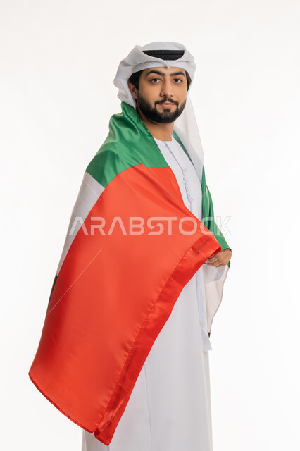 النظر إلى الكاميرا بإيماءات السعادة والسرور، الاحتفال بالأعياد والمناسبات الوطنية, الانتماء والاعتزاز بالهوية الإماراتية, اليوم الوطني الإماراتي 2 ديسمبر, بورتريه لرجل عربي خليجي إماراتي يرتدي الكندورة والغترة البيضاء يضع علم الإمارات على ظهره، خلفية بيضاء