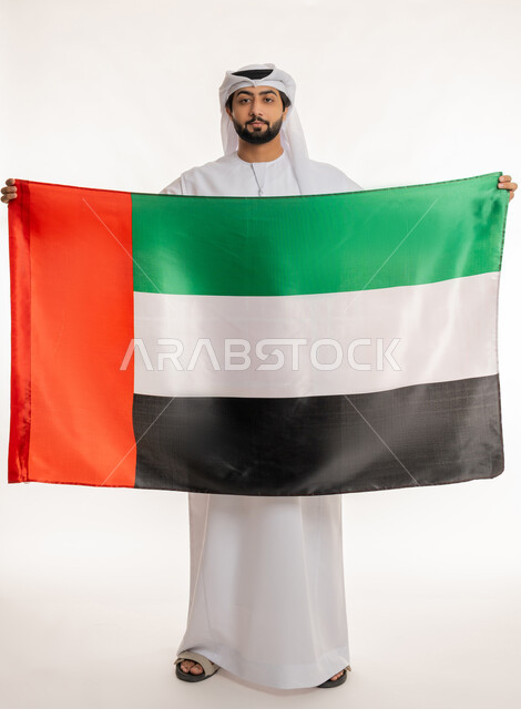 الاحتفال بالأعياد والمناسبات الوطنية, النظر إلى الكاميرا بتعابير الفخر والاعتزاز بالهوية الإماراتية, ذكرى اليوم الوطني الإماراتي 2 ديسمبر, بورتريه لرجل عربي خليجي إماراتي يرتدي الكندورة والغترة البيضاء يحمل علم الإمارات, الجسم بالطول الكامل، خلفية بيضاء