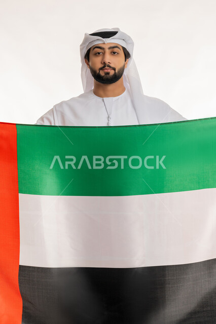النظر إلى الكاميرا بتعابير الفخر والاعتزاز بالهوية الإماراتية, الاحتفال بالأعياد والمناسبات الوطنية, ذكرى اليوم الوطني الإماراتي 2 ديسمبر, بورتريه لرجل عربي خليجي إماراتي يرتدي الكندورة والغترة البيضاء يحمل علم الإمارات، خلفية بيضاء