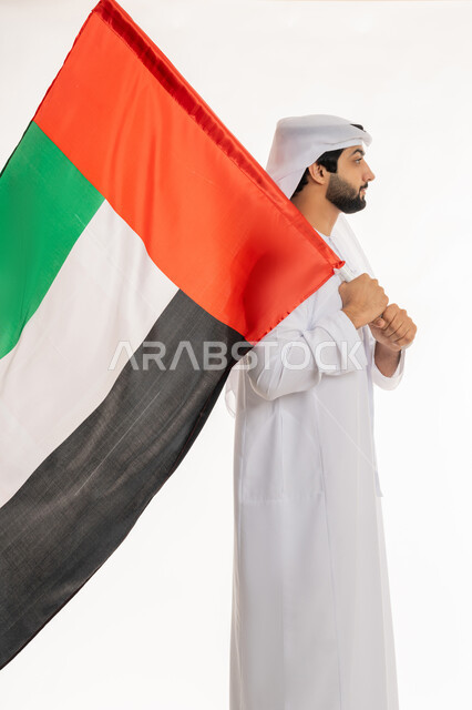 تعابير الانتماء والاعتزاز  بالهوية الإماراتية, الاحتفال بالأعياد والمناسبات الوطنية, مفهوم الحب والفخر بالوطن, ذكرى اليوم الوطني الإماراتي 2 ديسمبر, بورتريه من الجانب لرجل عربي خليجي إماراتي يرتدي الكندورة والغترة البيضاء يحمل علم الإمارات، خلفية بيضاء