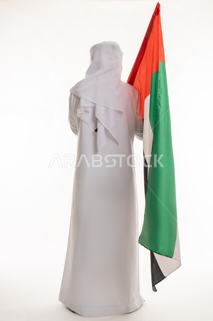 مفهوم الحب والفخر بالوطن, الاحتفال بالأعياد والمناسبات الوطنية, ذكرى اليوم الوطني الإماراتي 2 ديسمبر, بورتريه من الخلف لرجل عربي خليجي إماراتي يرتدي الكندورة والغترة البيضاء يحمل علم الإمارات, الجسم بالطول الكامل، خلفية بيضاء
