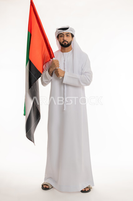 النظر إلى الكاميرا بتعابير الفخر والاعتزاز بالهوية الإماراتية, الاحتفال بالأعياد والمناسبات الوطنية, ذكرى اليوم الوطني الإماراتي 2 ديسمبر, بورتريه لرجل عربي خليجي إماراتي يرتدي الكندورة والغترة البيضاء يحمل علم الإمارات, الجسم بالطول الكامل، خلفية بيضاء