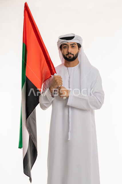 النظر إلى الكاميرا بتعابير الفخر والاعتزاز بالهوية الإماراتية, الاحتفال بالأعياد والمناسبات الوطنية, ذكرى اليوم الوطني الإماراتي 2 ديسمبر, بورتريه لرجل عربي خليجي إماراتي يرتدي الكندورة والغترة البيضاء يحمل علم الإمارات، خلفية بيضاء