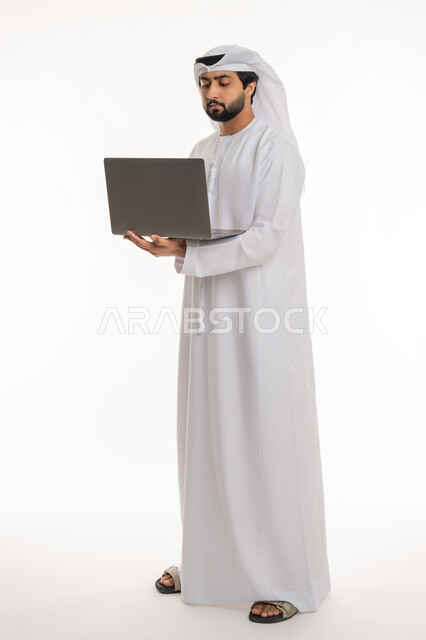تصفح مواقع التواصل الاجتماعي والعمل عن بعد, إيماءات التركيز في شاشة الكمبيوتر, استخدام الأجهزة التقنية الحديثة لإنجاز المهام والمشاريع, بورتريه لرجل عربي خليجي إماراتي يرتدي الكندورة والغترة البيضاء يعمل على الحاسوب المحمول, الجسم بالطول الكامل, خلفية بيضاء
