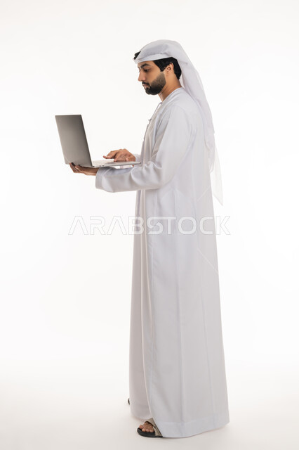استخدام الأجهزة التقنية الحديثة لإنجاز المهام والمشاريع, تصفح مواقع التواصل الاجتماعي والعمل عن بعد, إيماءات التركيز في شاشة الكمبيوتر, بورتريه لرجل عربي خليجي إماراتي يرتدي الكندورة والغترة البيضاء يعمل على الحاسوب المحمول, الجسم بالطول الكامل, خلفية بيضاء