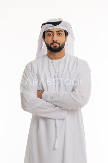 النظر إلى الكاميرا بإيماءات الحزم والجدية, مفهوم الأناقة والرجولة, الاهتمام بالمظهر الخارجي, بورتريه لرجل عربي خليجي إماراتي يرتدي الكندورة والغترة البيضاء يقف بأيدي متقاطعة بتعابير الثقة بالنفس, خلفية بيضاء