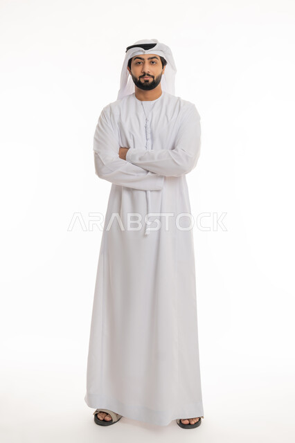 مفهوم الأناقة والرجولة, الاهتمام بالمظهر الخارجي, بورتريه لرجل عربي خليجي إماراتي يرتدي الكندورة والغترة البيضاء يقف بأيدي متقاطعة بتعابير الثقة بالنفس, النظر إلى الكاميرا بإيماءات الحزم والجدية, الجسم بالطول الكامل, خلفية بيضاء