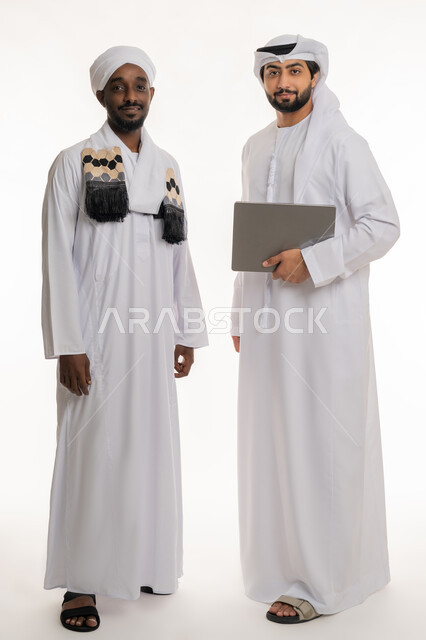 استخدام الأجهزة التقنية الحديثة والمتطورة, النظر إلى الكاميرا بإيماءات السعادة والسرور, قضاء الأوقات الممتعة مع الأصدقاء, بورتريه لشاب عربي خليجي إماراتي يرتدي الكندورة والغترة البيضاء يقف مع رجل سوداني يرتدي الجلابية والعمامة, الجسم بالطول الكامل, خلفية بيضاء