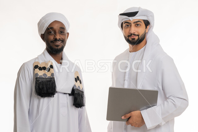 النظر إلى الكاميرا بإيماءات السعادة والسرور, قضاء الأوقات الممتعة مع الأصدقاء, استخدام الأجهزة التقنية الحديثة والمتطورة, بورتريه مقرب لشاب عربي خليجي إماراتي يرتدي الكندورة والغترة البيضاء يقف مع رجل سوداني يرتدي الجلابية والعمامة, خلفية بيضاء