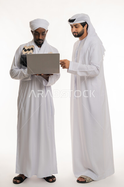 استخدام الأجهزة التقنية الحديثة والمتطورة, التعاون في إنجاز مهام العمل على الحاسوب المحمول, مناقشة الخطط والمشاريع, بورتريه لشاب عربي خليجي إماراتي يرتدي الكندورة والغترة البيضاء يعمل مع رجل سوداني يرتدي الجلابية والعمامة, الجسم بالطول الكامل, خلفية بيضاء