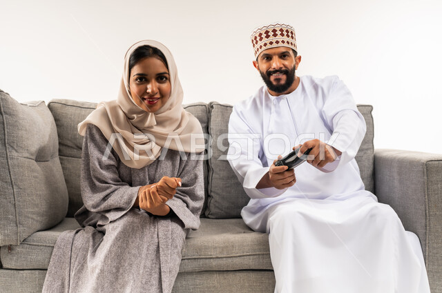 الاستمتاع والتسلية في العطلة، نشاط ترفيهي منزلي، تعابير الحزن للخسارة، استخدام جهاز تكنولوجي حديث، لعب ألعاب الفيديو في المنزل، بورتريه لرجل عربي خليجي عماني يرتدي الدشداشة والكمة يجلس على الكنبة في غرفة المعيشة بجانب زوجته يحمل بيده عصا التحكم، خلفية بيضاء