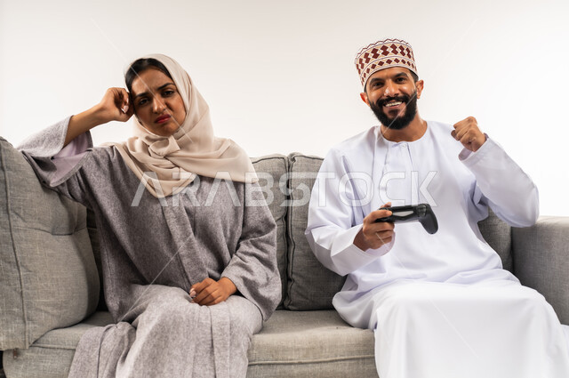 نشاط ترفيهي منزلي، تعابير الحزن للخسارة، استخدام جهاز تكنولوجي حديث، لعب ألعاب الفيديو في المنزل، بورتريه لرجل عربي خليجي عماني يرتدي الدشداشة والكمة يجلس على الكنبة بجانب زوجته يحمل بيده عصا التحكم، امرأة تضع يدها على رأسها بايماءات الاحباط، خلفية بيضاء