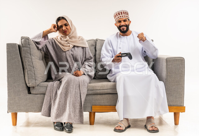 تعابير الحزن للخسارة، نشاط ترفيهي منزلي، استخدام جهاز تكنولوجي حديث، لعب ألعاب الفيديو في المنزل، بورتريه لرجل عربي خليجي عماني يرتدي الدشداشة والكمة يجلس على الكنبة بجانب زوجته يحمل بيده عصا التحكم، امرأة تضع يدها على رأسها بايماءات الاحباط، خلفية بيضاء
