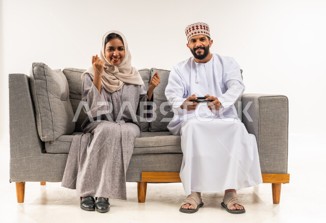 استخدام جهاز تكنولوجي حديث، لعب ألعاب الفيديو في المنزل، نشاط ترفيهي منزلي، بورتريه لرجل عربي خليجي عماني يرتدي الدشداشة والكمة يجلس على كنبة مريحة في غرفة المعيشة بجانب زوجته يحمل بيده عصا التحكم، امرأة ترفع يديها بايماءات النصر، خلفية بيضاء