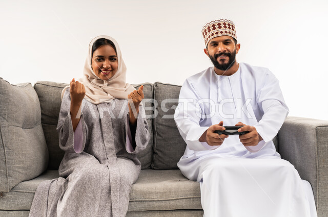 لعب ألعاب الفيديو في المنزل، استخدام الأجهزة التكنولوجيا الحديثة، نشاط ترفيهي منزلي، بورتريه لرجل عربي خليجي عماني يرتدي الدشداشة والكمة يجلس على كنبة مريحة في غرفة المعيشة بجانب زوجته يحمل بيده عصا التحكم، امرأة ترفع يديها بايماءات النصر، خلفية بيضاء
