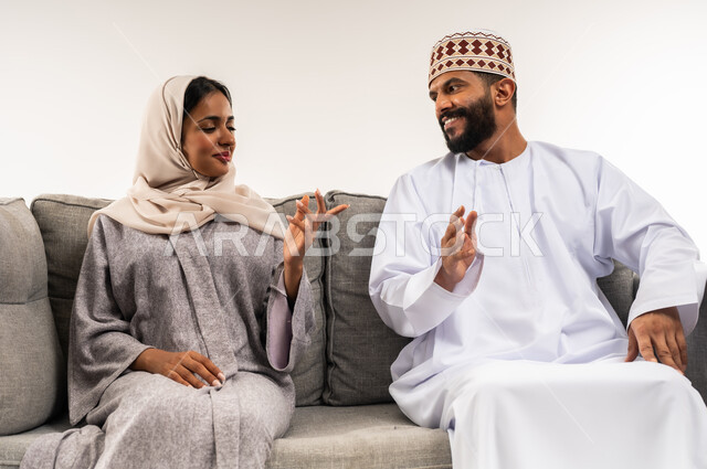 تبادل الاحاديث والاخبار بين افراد العائلة، المحافظة على الترابط الأسري، مفهوم الأمان والإستقرار العائلي، بورتريه لزوجين عربيي خليجيين عمانيين يجلسان على اريكة مريحة في غرفة المعيشة، خلفية بيضاء