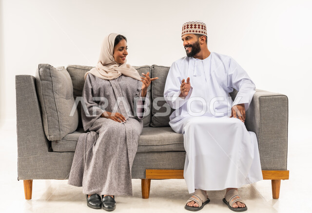 تبادل الاحاديث والاخبار بين افراد العائلة، المحافظة على الترابط الأسري، مفهوم الأمان والإستقرار العائلي، بورتريه لزوجين عربيي خليجيين عمانيين يجلسان على اريكة مريحة في غرفة المعيشة، خلفية بيضاء