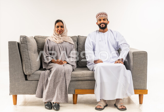 النظرالى الكاميرا بايماءات السرور والسعادة، المحافظة على الترابط الأسري، مفهوم الأمان والإستقرار العائلي، بورتريه لزوجين عربيي خليجيين عمانيين يجلسان على اريكة مريحة في غرفةالمعيشة، خلفية بيضاء