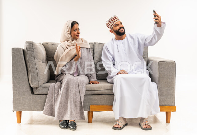 Greeting and peace gestures, concept of family security and stability, making an online video call, documenting happy family moments, portrait of an Arab Gulf Omani couple taking a souvenir photo using a mobile phone, maintaining family ties, white background
