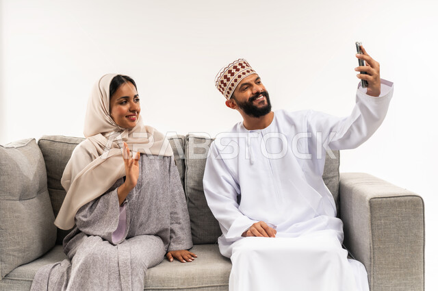 ايماءات القاء التحية والسلام، مفهوم الأمان والإستقرار العائلي، إجراء مكالمة فيديو أونلاين، توثيق اللحظات العائلية السعيدة، بورتريه لزوجين عربيي خليجيين عمانيين يلتقطان صورة تذكارية من خلال الهاتف المحمول، المحافظة على الترابط الأسري، خلفية بيضاء