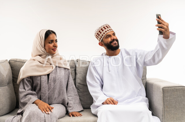 المحافظة على الترابط الأسري، مفهوم الأمان والإستقرار العائلي، إجراء مكالمة فيديو أونلاين، توثيق اللحظات العائلية السعيدة، بورتريه لزوجين عربيي خليجيين عمانيين يلتقطان صورة تذكارية من خلال الهاتف المحمول، خلفية بيضاء