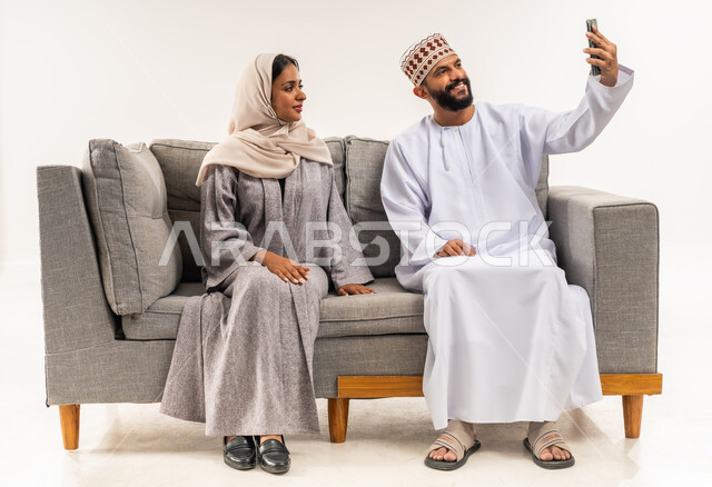 إجراء مكالمة فيديو أونلاين، المحافظة على الترابط الأسري، مفهوم الأمان والإستقرار العائلي، توثيق اللحظات العائلية السعيدة، بورتريه لزوجين عربيي خليجيين عمانيين يلتقطان صورة تذكارية من خلال الهاتف المحمول، خلفية بيضاء