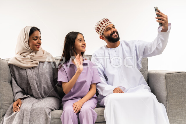 مفهوم الأمان والإستقرار العائلي، إجراء مكالمة فيديو أونلاين، توثيق اللحظات العائلية السعيدة، ايماءات القاء التحية والسلام، بورتريه لعائلة عربية خليجية عمانية تلتقط صورة تذكارية من خلال الهاتف المحمول، المحافظة على الترابط الأسري، خلفية بيضاء