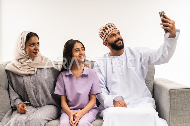 إجراء مكالمة فيديو أونلاين، توثيق اللحظات العائلية السعيدة، بورتريه لعائلة عربية خليجية عمانية تلتقط صورة تذكارية من خلال الهاتف المحمول، مفهوم الأمان والإستقرار العائلي، المحافظة على الترابط الأسري، خلفية بيضاء