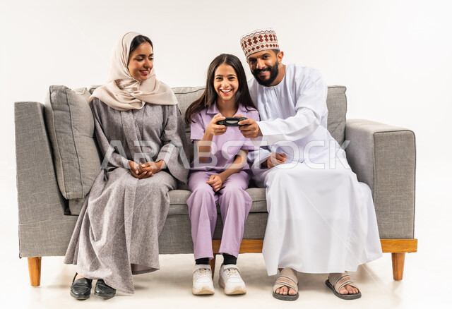 مشاركة المرح مع الاسرة، لعب العاب الفيديو الترفيهية، استخدام التكنولوجيا الحديثة، بورتريه لفتاة عربية خليجية عمانية تجلس على اريكة مريحة في غرفة المعيشة برفقة والديها تحمل بيدها عصا التحكم، قضاء وقت ممتع مع الابناء، أجواء عائلية سعيدة، خلفية بيضاء