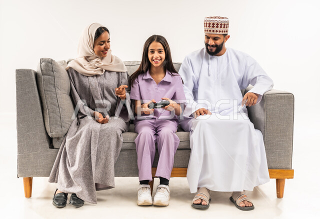 لعب العاب الفيديو الترفيهية، استخدام التكنولوجيا الحديثة، بورتريه لفتاة عربية خليجية عمانية تجلس على اريكة مريحة في غرفة المعيشة برفقة والديها تحمل بيدها عصا التحكم، قضاء وقت ممتع مع الابناء، أجواء عائلية سعيدة، مشاركة المرح مع الاسرة، خلفية بيضاء