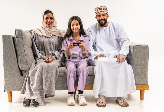 Using modern technology, playing entertaining video games, portrait of an Arab Gulf Omani girl sitting on a comfortable sofa in the living room with her parents holding a joystick, spending quality time with children, happy family atmosphere, sharing fun with family, white background