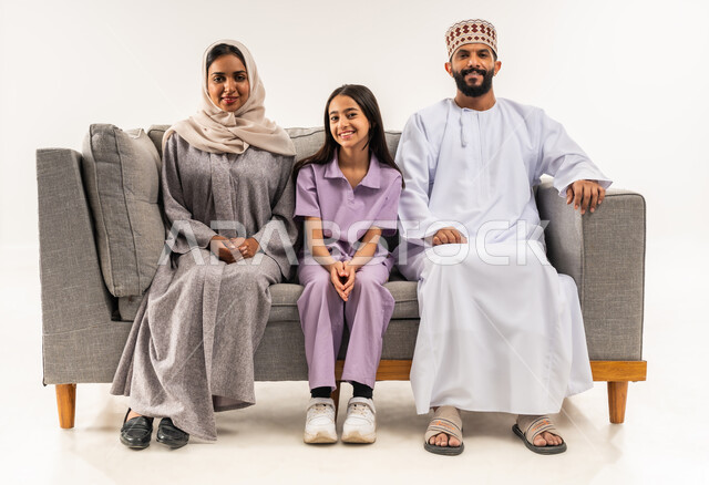النظر الى الكاميرا بايماءات السرور والسعادة، بورتريه لزوجين عربيين خليجيين عمانيين يجلسان على اريكة مريحة في غرفة المعيشة برفقة ابنتهما، قضاء وقت ممتع مع الابناء، أجواء عائلية سعيدة، مشاركة المرح مع الاسرة، خلفية بيضاء