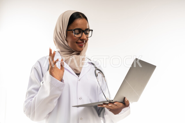 تقديم الاستشارة عبر الانترنت، اعداد التقارير الطبية، بورتريه مقرب لطبيبة عربية خليجية عمانية محجبة ترتدي المعطف الطبي الأبيض تضع سماعة الفحص تحمل الحاسوب المحمول ترفع يدها بايماءات التحية, شابة سعودية تعمل بمجال الرعاية الصحية، خلفية بيضاء