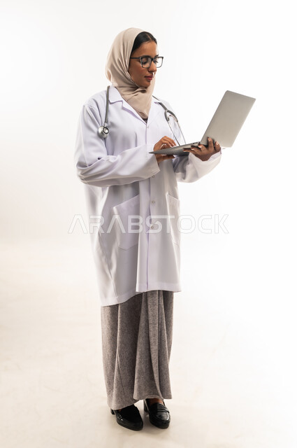 Preparing medical reports, providing online consultation, portrait of a veiled Gulf Arab Omani female doctor wearing a white medical coat, holding a stethoscope and holding a laptop in her hand, a young Saudi woman working in the health care field, full body image, white background