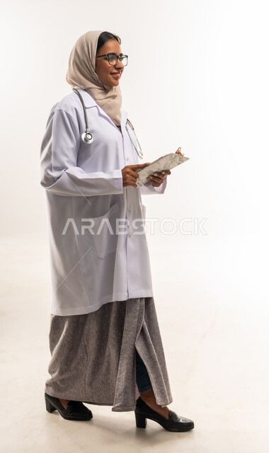 ايماءات المشي والذهاب لمكان ما، الطب والرعاية الصحية، تدوين الملاحظات والبيانات، بورتريه من الجانب لطبيبة عربية خليجية عمانية محجبة ترتدي المعطف الطبي تضع سماعة الفحص تحمل بيدها حامل الاوراق, شابة سعودية تقوم بتقديم المساعدة الطبية، صورة لكامل الجسم، خلفية بيضاء