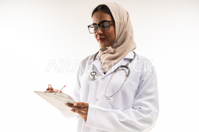 العمل في المراكز والمستشفيات، الطب والرعاية الصحية، تدوين الملاحظات والبيانات، بورتريه مقرب لطبيبة عربية خليجية عمانية محجبة ترتدي المعطف الطبي الأبيض تضع سماعة الفحص الطبية تحمل بيدها حامل الاوراق, شابة سعودية تقوم بتقديم المساعدة الطبية، خلفية بيضاء