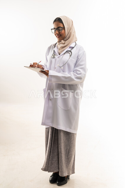 تدوين الملاحظات والبيانات، الطب والرعاية الصحية، العمل في المراكز والمستشفيات، بورتريه لطبيبة عربية خليجية عمانية محجبة ترتدي المعطف الطبي الأبيض تضع سماعة الفحص الطبية تحمل بيدها حامل الاوراق, شابة سعودية تقوم بتقديم المساعدة الطبية، صورة لكامل الجسم، خلفية بيضاء