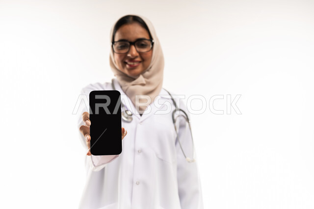 Medicine and healthcare, blank black screen display, working in centers and hospitals, close-up portrait of veiled Gulf Arab Omani female doctor wearing white medical coat wearing stethoscope holding mobile phone in hand, young Saudi woman providing medical assistance, white background